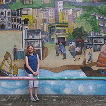 Emma Lenz in front of a mural with the sea, ships, houses, and people walking